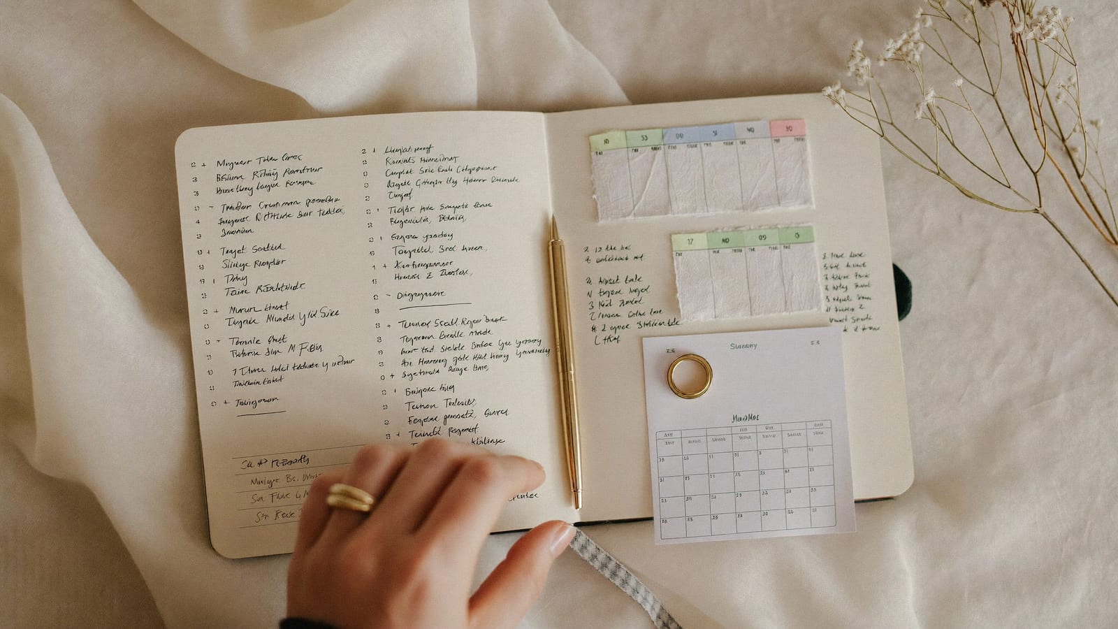 Agendă de organizare a nunții deschisă, cu listă bifată, pix auriu, calendar și flori albe uscate pe o față de masă crem